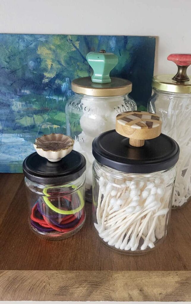 use glass jars in the bathroom as DIY bathroom storage by attaching knobs to the tops of old food jars and storing qtips, hairbands, and cotton balls